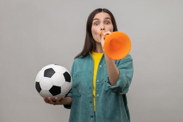 Aufgeregte Frau, die in Horn bläst und einen Fußball hält, der den Sieg der Lieblingsfußballmannschaft feiert