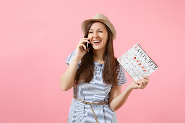 Aufgeregte Frau, die mit dem Handy spricht, angenehme Gespräche führt, den Periodenkalender hält, um die Menstruationstage einzeln auf rosafarbenem Hintergrund zu überprüfen. Medizin, Gesundheitskonzept. Platz kopieren.