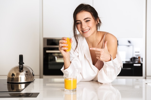 aufgeregte Frau, die Seidenkleidung trägt, die frischen Saft mit Orange macht, während des Frühstücks in der Küche zu Hause