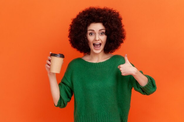 Aufgeregte Frau mit Afro-Frisur, die einen grünen, lässigen Pullover trägt, der Kaffee zum Mitnehmen hält und den Daumen nach oben zeigt, empfiehlt ein Kaffeehaus. Studioaufnahme im Innenbereich isoliert auf orangefarbenem Hintergrund