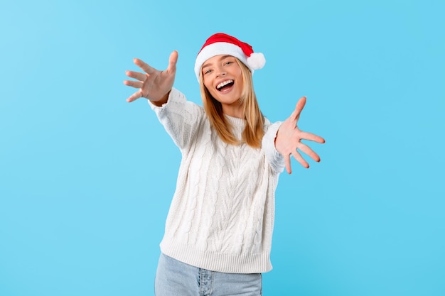 Aufgeregte Frau mit Weihnachtsmannshut reicht der Kamera auf blauem Hintergrund entgegen
