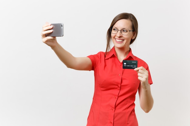 Aufgeregte Geschäftsfrau im roten Hemd, die selfie auf dem Handy mit Kreditkarte, bargeldlosem Geld einzeln auf weißem Hintergrund erschossen. Bildungsunterricht im Hochschulkonzept.