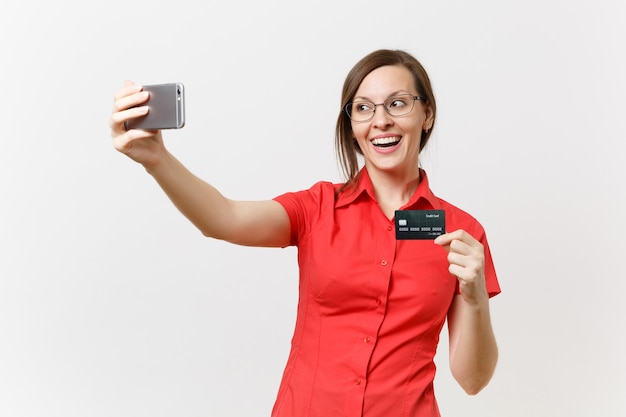 Aufgeregte Geschäftsfrau im roten Hemd, die selfie auf dem Handy mit Kreditkarte, bargeldlosem Geld einzeln auf weißem Hintergrund erschossen. Bildungsunterricht im Hochschulkonzept.