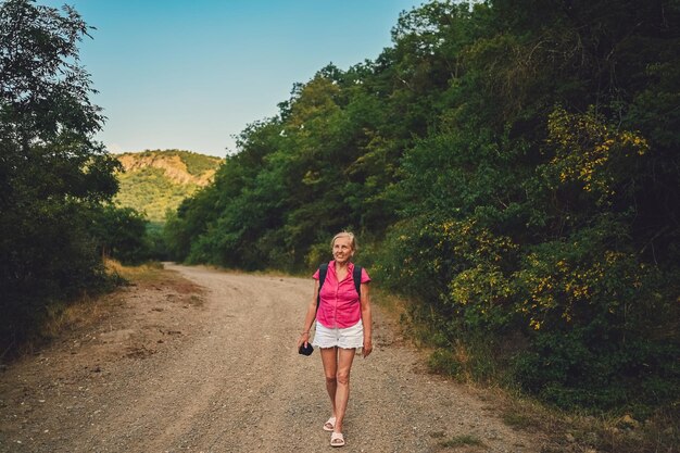 Aufgeregte, glückliche, ältere Frau, Rucksack-Touristin, die im Sommer in der Waldstraße im Freien bei Sonnenuntergang spazieren geht