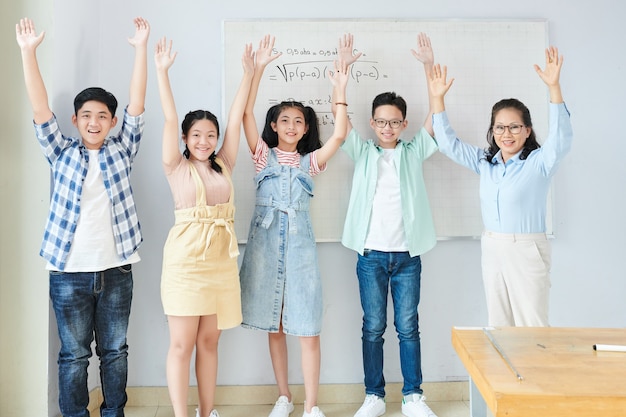 Aufgeregte glückliche asiatische Schüler und ihr Lehrer für Naturwissenschaften, die mit Gleichungen vor dem Whiteboard stehen, die Arme heben und das Beenden eines schwierigen Mathe-Themas feiern