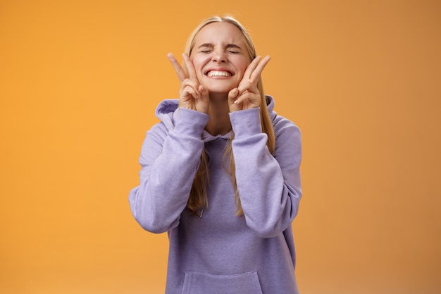 Aufgeregte glückliche charmante freundliche freundliche blonde Freundin, die Spaß hat, perfekte Nachrichten zu feiern beste Punktzahl erhalten Stipendium lächelnd amüsiert sorglos schließen Augen zeigen Friedenssieg Zeichen, orange Hintergrund.