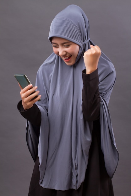 Aufgeregte, glückliche muslimische Frau, die Smartphone, drahtloses Internetgerät verwendet