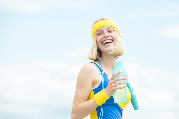 Aufgeregte, glückliche Sportlerin, die selbstbewusst in die Kamera schaut, während sie eine Flasche Wasser in gelbblauer Kleidung hält, die auf der Straße trainiert