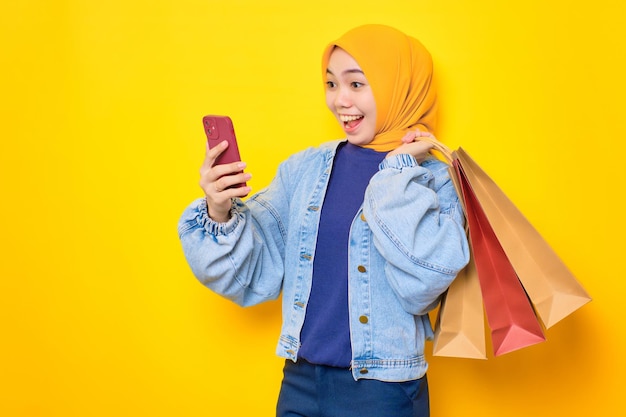 Aufgeregte junge Asiatin in Jeansjacke mit Handy für Online-Shopping mit Einkaufstüten isoliert auf gelbem Hintergrund