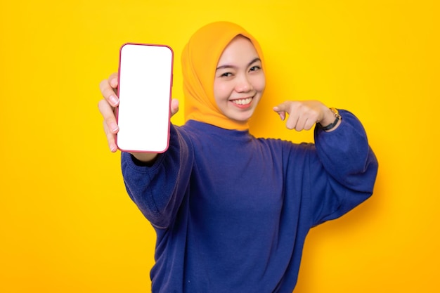 Aufgeregte junge asiatische Muslimin in einem lässigen Pullover, die ein Handy mit leerem Bildschirm zeigt, das eine mobile App isoliert über gelbem Hintergrund empfiehlt