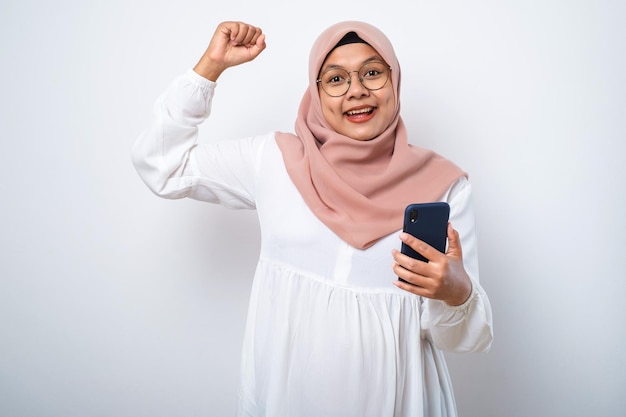 Aufgeregte junge asiatische muslimische Frau, die ein Mobiltelefon benutzt und den Erfolg feiert, gute Nachrichten zu bekommen