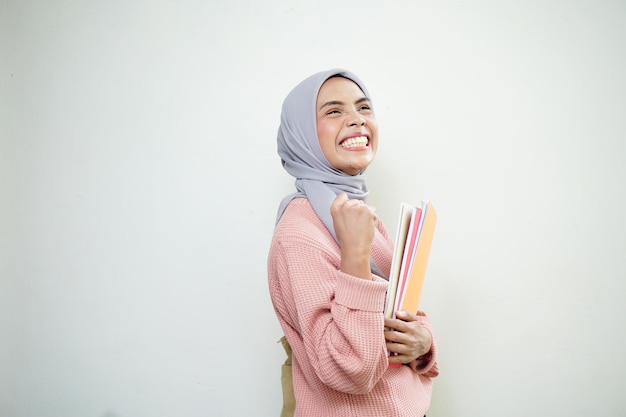Aufgeregte junge asiatische muslimische Studentin in rosa Pullover mit Tasche, die ein Buch hält