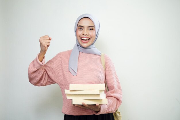 Aufgeregte junge asiatische muslimische Studentin in rosafarbenem Pullover mit Tasche und Haltebuch