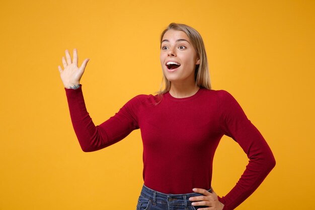 Aufgeregte junge blonde Frau Mädchen in Freizeitkleidung posiert isoliert auf gelb-orangefarbenem Hintergrund im Studio. People-Lifestyle-Konzept. Mock-up-Kopienbereich. Winken und Grüßen mit der Hand, wie jemand bemerkt.