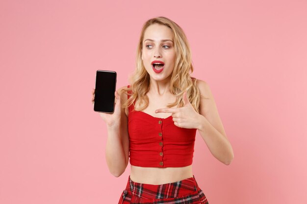 Aufgeregte junge blonde Frau Mädchen in roter sexy Kleidung posiert isoliert auf pastellrosa Hintergrund. People-Lifestyle-Konzept. Mock-up-Kopienbereich. Zeigen Sie mit dem Zeigefinger auf das Mobiltelefon mit leerem, leerem Bildschirm.