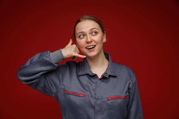 Aufgeregte junge blonde Ingenieurin in Uniform, die nach oben schaut, um Anrufgeste zu tun