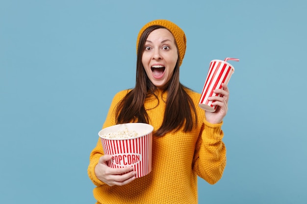 Aufgeregte junge brünette Frau in Pullovermütze posiert isoliert auf blauem Hintergrund. Menschen Emotionen im Kino-Lifestyle-Konzept. Mock-up-Kopienbereich. Anschauen von Filmen hält einen Eimer mit Popcorn und eine Tasse Soda.