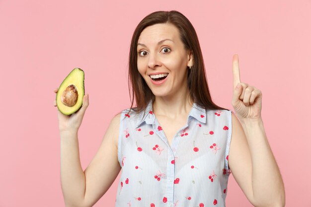 Aufgeregte junge Frau, die den Zeigefinger mit einer neuen Idee nach oben zeigt, hält die Hälfte der frischen reifen Avocado-Frucht isoliert auf rosa Pastellhintergrund. Menschen lebendiger Lebensstil entspannen Urlaubskonzept. Mock-up-Kopienbereich.