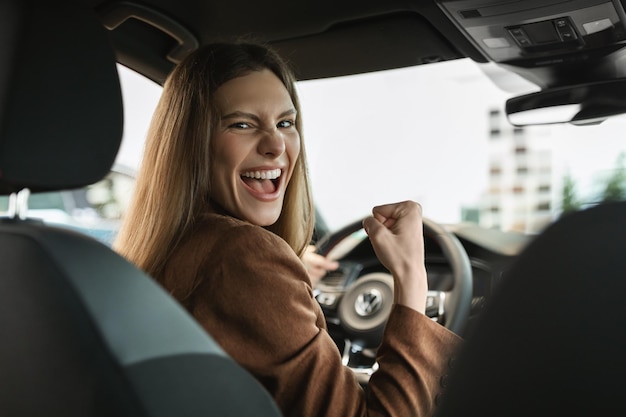 Aufgeregte junge Frau feiert erfolgreichen Autokauf und gestikuliert ja, während sie im Auto sitzt