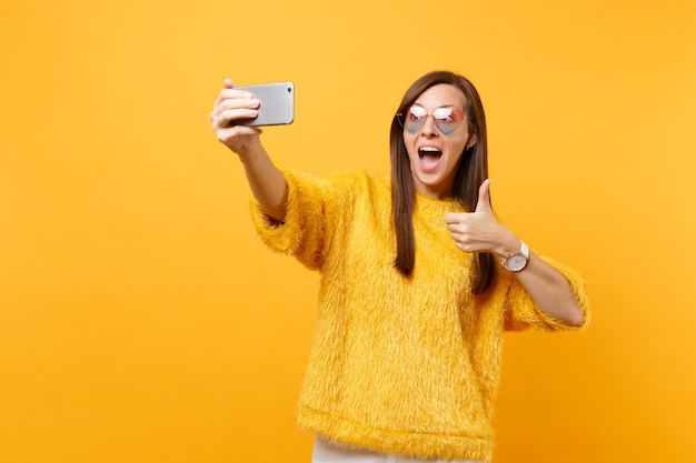 Aufgeregte junge Frau im Pelzpullover, Herzbrille zeigt Daumen nach oben und macht Selfie-Aufnahmen auf dem Handy einzeln auf hellgelbem Hintergrund. Menschen aufrichtige Emotionen, Lebensstil. Werbefläche.