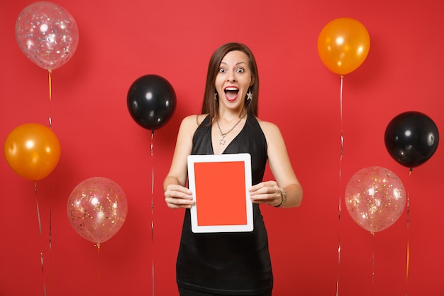Aufgeregte junge Frau im schwarzen Kleid feiert, Tablet-PC-Computer mit leerem schwarzem leerem Bildschirm auf leuchtend roten Hintergrundluftballons halten. Frohes neues Jahr, Geburtstagsmodell Urlaub Party Konzept.