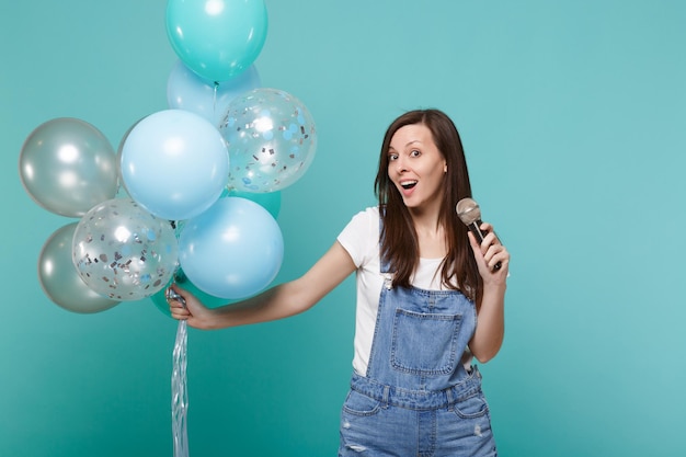Aufgeregte junge Frau in Denim-Kleidung singt ein Lied im Mikrofon, feiert und hält bunte Luftballons einzeln auf blau-türkisem Wandhintergrund. Geburtstagsfeier, Menschen Emotionen Konzept.