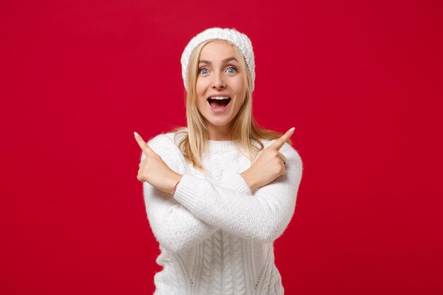 Aufgeregte junge Frau in weißem Pulloverhut isoliert auf rotem Hintergrund im Studio. Gesundes Mode-Lifestyle-Konzept der kalten Jahreszeit. Mock-up-Kopienbereich. Zeigefinger zur Seite zeigen, Hände gekreuzt halten.
