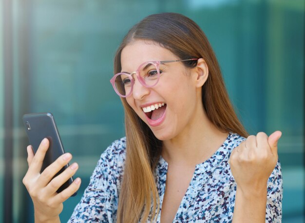 Aufgeregte junge Frau mit Brille, die ihr Smartphone im Freien überprüft