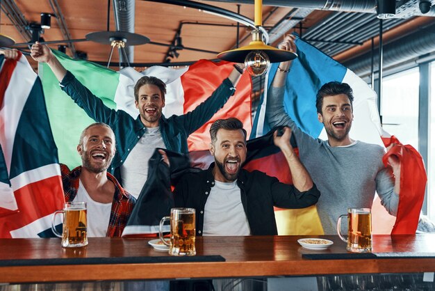 Aufgeregte junge Männer, die mit internationalen Flaggen bedeckt sind, trinken Bier und schauen sich Sportspiele an, während sie in der Kneipe sitzen