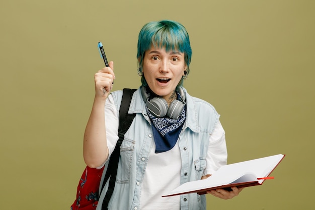 Aufgeregte junge Studentin mit Kopfhörern und Bandana am Hals und Rucksack mit offenem Notizbuch, das in die Kamera blickt, die mit einem Stift isoliert auf olivgrünem Hintergrund nach oben zeigt