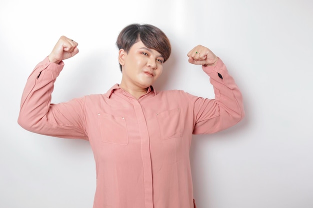 Aufgeregte kurzhaarige asiatische Frau, die ein rosafarbenes Hemd trägt, das eine starke Geste zeigt, indem sie ihre Arme und Muskeln stolz lächelt