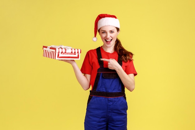 Aufgeregte Lieferfrau, die eingewickelte Geschenkbox auf der Handfläche hält und mit dem Finger auf das Geschenk zeigt, blaue Overalls und Weihnachtsmannmütze trägt. Studioaufnahme im Innenbereich isoliert auf gelbem Hintergrund.