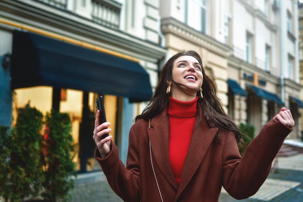 Foto aufgeregte modische frau feiert gute nachrichten und hält das telefon in der abendstraße