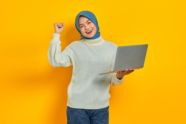 Aufgeregte schöne asiatische Frau im weißen Pullover, die einen Laptop hält und den Sieg isoliert über gelbem Hintergrund feiert Menschen religiöses Lebensstilkonzept