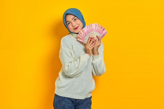 Aufgeregte schöne asiatische Muslimin in weißem Pullover hält einen Geldfächer in Rupiah-Banknoten und betrachtet den Kopienraum isoliert auf gelbem Hintergrund Religiöses Lifestyle-Konzept der Menschen