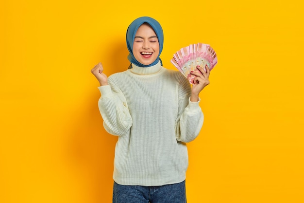 Aufgeregte schöne asiatische muslimische Frau in weißem Pullover, die Bargeld in indonesischen Rupiah-Banknoten hält und das Glück isoliert über gelbem Hintergrund feiert Religiöses Lifestyle-Konzept der Menschen