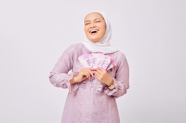 Aufgeregte schöne junge asiatische Muslimin mit Hijab und lila Kleid, die Bargeld in Rupiah-Banknoten hält, während sie isoliert auf weißem Studiohintergrund in die Kamera schaut