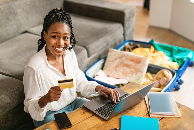 Aufgeregte schwarze Frau, die Laptop benutzt und Tour oder Hotel online bucht, mit Kreditkarte, die sich vorbereitet