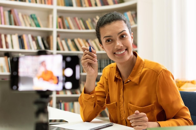 Aufgeregte Tutorin gemischter Rassen, die einen Online-Vortrag hält und mit einer Smartphone-Kamera spricht, die einen Videoanruf in der Bibliothek tätigt