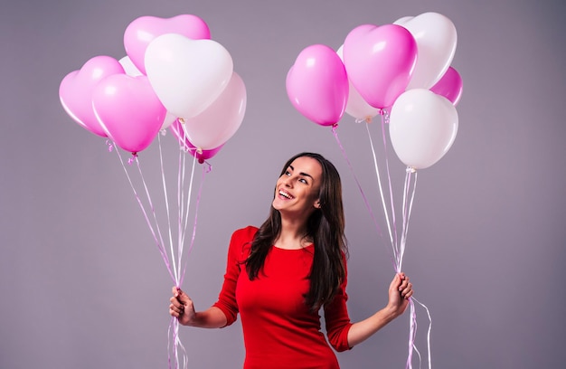 Aufgeregte und glückliche, elegante junge Frau in Rot hält in den Händen viele bunte Ballons, die im Studio isoliert sind