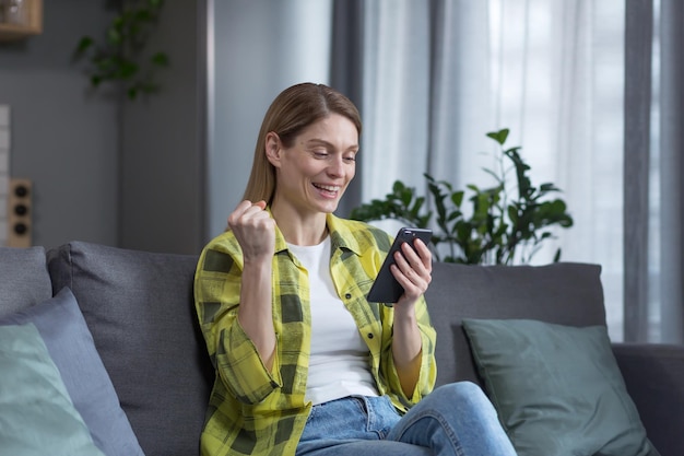 Aufgeregte und glückliche Frau, die sich den Telefonbildschirm anschaut, gewinnt und gute Nachrichten Siegesgeste Frau, die zu Hause auf dem Sofa sitzt