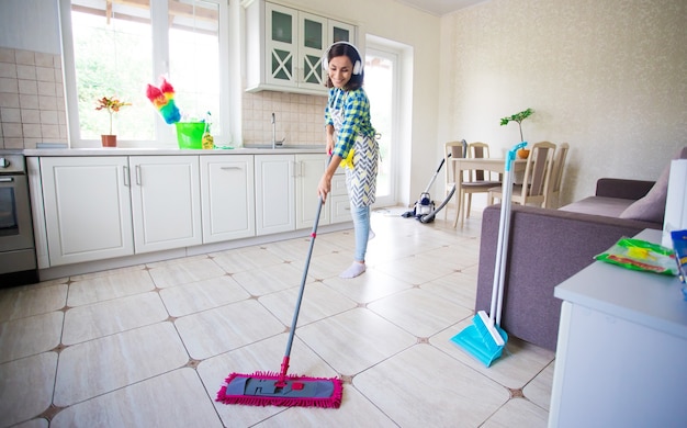 Aufgeregte und glückliche schöne junge Frau in einer Schürze putzt den Boden in ihrer Küche zu Hause und tanzt