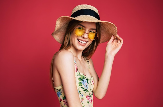 Aufgeregte und glückliche schöne junge Frau in Sonnenbrille, Kleid und Sommerhut hat Spaß beim Posieren auf rosa Hintergrund