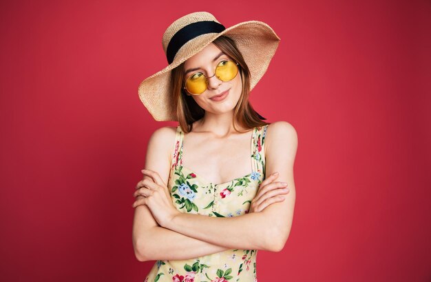 Aufgeregte und glückliche schöne junge Frau in Sonnenbrille, Kleid und Sommerhut hat Spaß beim Posieren auf rosa Hintergrund