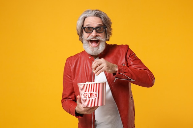 Aufgeregter älterer grauhaariger bärtiger Mann mit Schnurrbart in Lederjacke, 3D-Brille isoliert auf gelb-orangefarbenem Hintergrund. Menschen Emotionen im Kinokonzept. Film ansehen, Eimer Popcorn halten.