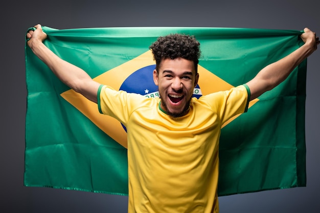 Aufgeregter afroamerikanischer Fußballfan schreit und hält brasilianische Flagge auf Grau