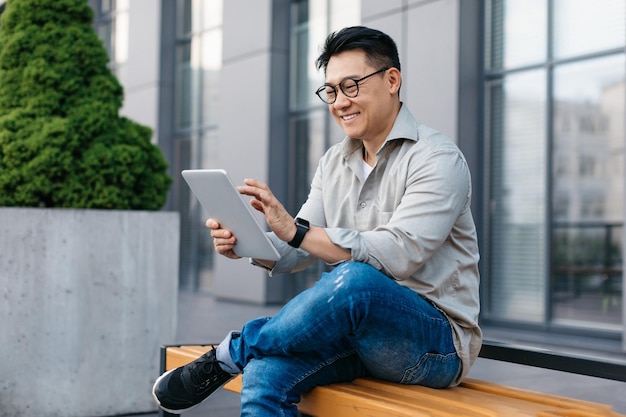 Aufgeregter asiatischer Geschäftsmann, der Tablets verwendet, um E-Mails zu checken oder mit Kunden zu chatten, sitzt auf einer Bank in der Nähe des Bürogebäudes