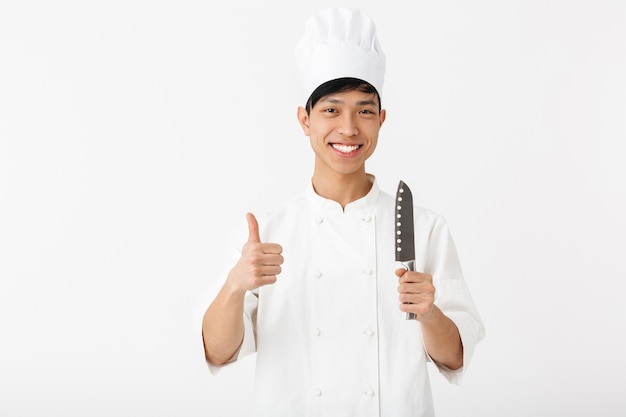 Aufgeregter asiatischer Koch, der Uniform trägt, die isoliert über weißer Wand steht und Küchenausstattung zeigt
