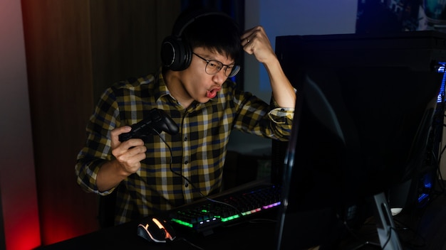 Aufgeregter asiatischer Mann pro Spieler, der am Tisch sitzt, spielt und gewinnt in Online-Videospielen auf einem Computer und Smartphone, Technologie-Gaming-Cyber- oder E-Sport-Meisterschaftskonzept