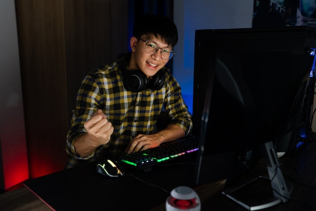 Aufgeregter asiatischer Mann pro Spieler, der am Tisch sitzt, spielt und gewinnt in Online-Videospielen auf einem Computer und Smartphone, Technologie-Gaming-Cyber- oder E-Sport-Meisterschaftskonzept.